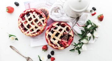 An overhead shot of strawberry and blackberry pies next to a white coffee pot and a bunch of flowers