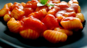 closeup of gnocchi covered in marinara sauce with a few green leaves of basil on a dark plate