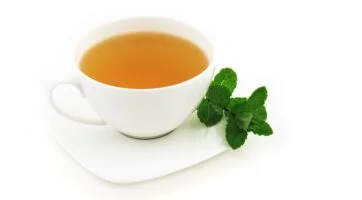 picture of peppermint tea in a white tea cup, on a white saucer, with green mint leaves on the sauce with a white background.